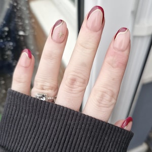 May include: Close-up of a hand with professionally manicured nails. The nails feature a clear base with a burgundy tip and a gold accent line. One nail has a gold star design. The hand is wearing a ring with a square-cut stone.