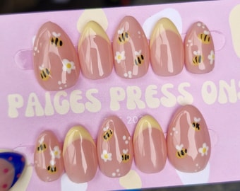 Puede incluir: Un juego de diez uñas postizas en forma de almendra. Las uñas son de color rosa claro con una punta francesa amarilla. Cada uña está decorada con una abeja y pequeñas flores blancas. El texto "PAIGES PRESS ONS" está impreso en el fondo rosa.