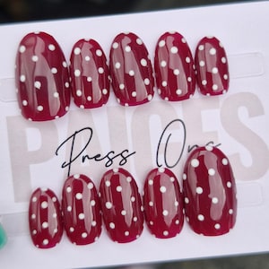 May include: A set of deep red press-on nails featuring white polka dots. The oval-shaped nails are displayed on a white card with the text "Press One". A teal object is visible in the corner. The nails are designed for easy application.