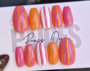 Puede incluir: Un conjunto de uñas postizas con varios diseños. Algunas uñas son de color rosa, naranja o blanco liso con rayas rosas. Otras tienen rayas verticales rosas y naranjas. La tarjeta dice "Press On".