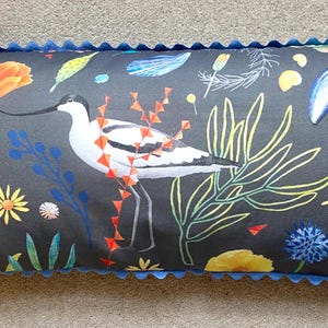 Puede incluir: Cojín rectangular con fondo gris oscuro que presenta una ilustración de un ave, flores y conchas marinas. El cojín tiene un borde festoneado azul. El ave tiene un pico largo y está rodeada de elementos botánicos coloridos.