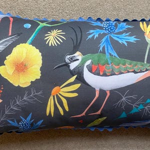 Puede incluir: Cojín rectangular con un fondo gris oscuro que presenta un diseño floral y de aves colorido. El ave tiene marcas verdes, blancas y naranjas. El cojín tiene un borde festoneado azul.