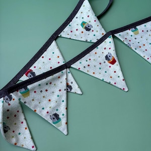 May include: A bunting with triangular flags. Each flag has a cream background with a pattern of hedgehogs in cups and small flowers. The flags are trimmed with dark grey and strung together with a grey ribbon. The bunting is suitable for a child's room.