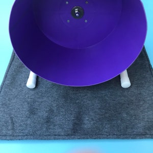 May include: A purple hamster wheel with white plastic supports sits on a grey fabric mat. The wheel is designed for small pets to exercise.