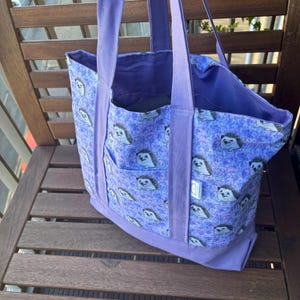 K&ouml;nnte beinhalten: Eine lilafarbene Einkaufstasche mit einem Igel-Druck. Die Tasche hat zwei gro&szlig;e Taschen auf der Vorderseite und zwei Henkel.