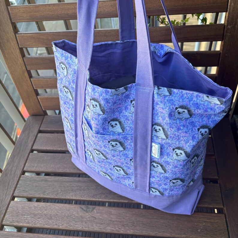 K&ouml;nnte beinhalten: Eine lilafarbene Einkaufstasche mit einem Igel-Druck. Die Tasche hat zwei gro&szlig;e Taschen auf der Vorderseite und zwei Henkel.