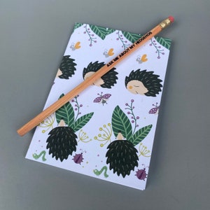 May include: A white notepad with a pencil on top. The notepad has a repeating pattern of hedgehogs, leaves, and bugs. The pencil has the text "Ask Me About My Hedgehog" printed on it.