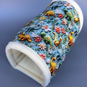 May include: A cylindrical fabric tunnel featuring a light blue background with a repeating pattern of colourful dinosaurs in shades of yellow, green, and blue, interspersed with various pink, white, and orange flowers and green foliage, finished with soft white trim.