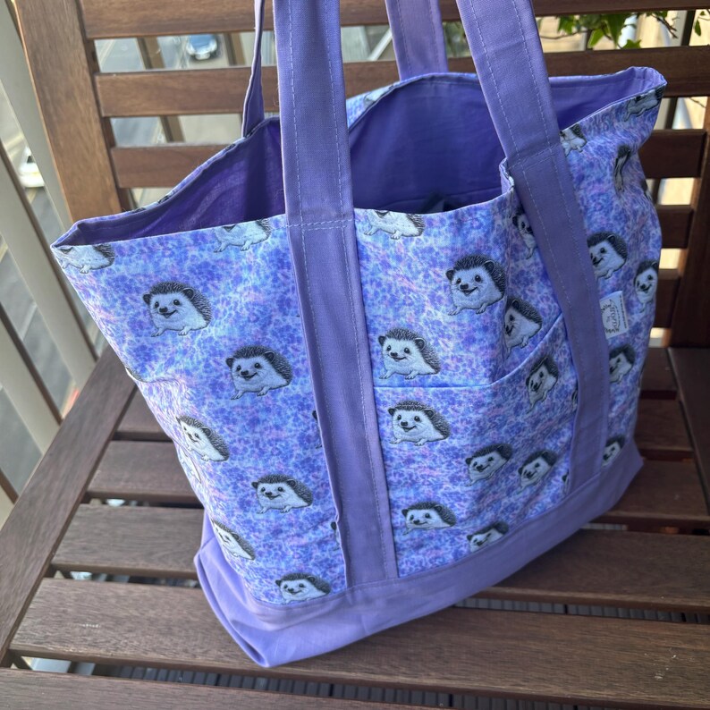 K&ouml;nnte beinhalten: Eine lilafarbene Einkaufstasche mit einem Muster aus Igeln auf einem helllilafarbenen Hintergrund. Die Tasche hat zwei lange lilafarbene Henkel.