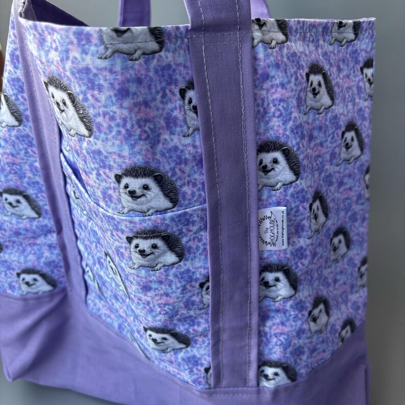 K&ouml;nnte beinhalten: Lilafarbene Einkaufstasche mit einem Batik-Muster mit Igeln. Die Tasche hat eine gro&szlig;e Tasche auf der Vorderseite und ein Etikett mit der Aufschrift "The Hedgehog House".