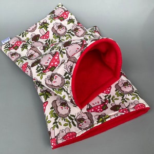 May include: A cream-colored fabric hedgehog-themed pet bed with a red fleece lining. The bed is shaped like a tunnel with a rounded opening.