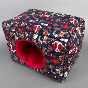 May include: A square pet bed with a navy blue patterned exterior, decorated with Christmas motifs such as Santa hats, reindeer, and presents. The interior is a bright red, with a circular entrance.