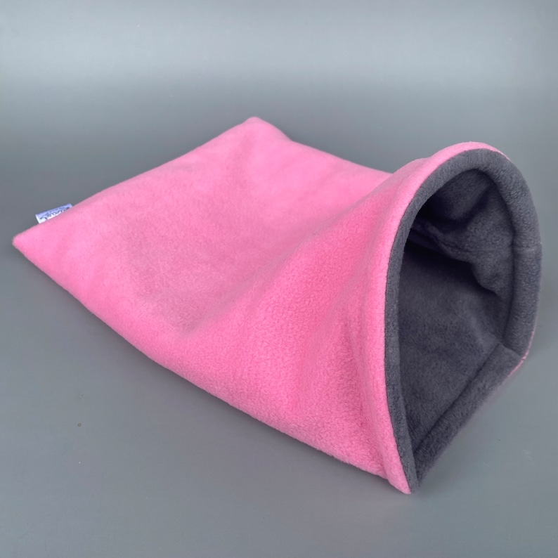 May include: Pink and gray fleece pet bed shaped like a tunnel. The bed is made of soft, plush fleece and has a cozy, enclosed design.