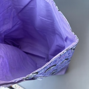 K&ouml;nnte beinhalten: Lilafarbene Stofftasche mit Rei&szlig;verschluss. Das Innere der Tasche ist mit einem einfarbigen lila Stoff gef&uuml;ttert. Die Au&szlig;enseite der Tasche hat ein Muster aus kleinen wei&szlig;en Pinguinen auf lila Hintergrund.