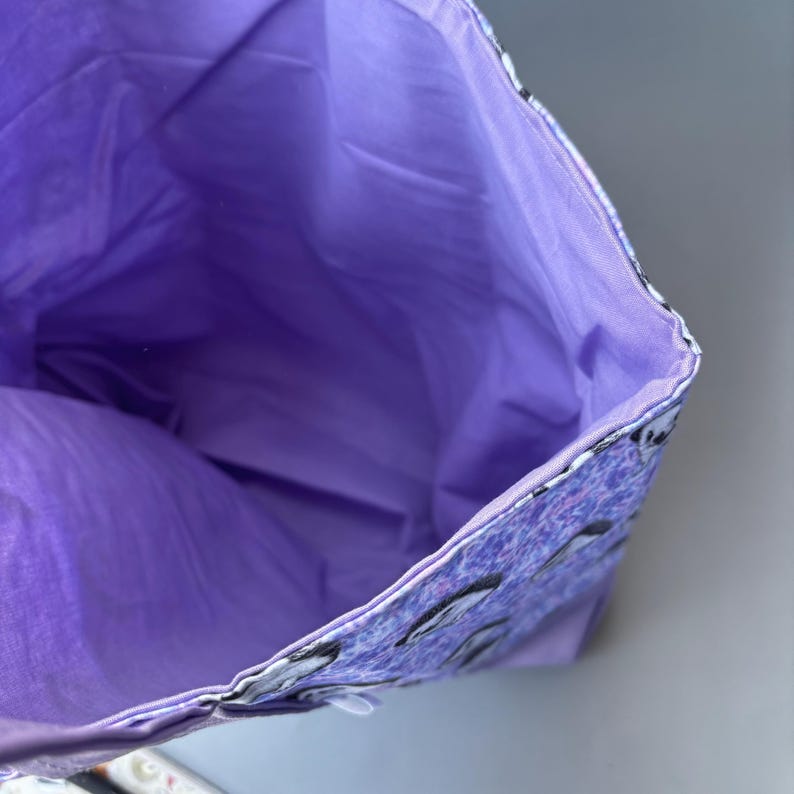 K&ouml;nnte beinhalten: Lilafarbene Stofftasche mit Rei&szlig;verschluss. Das Innere der Tasche ist mit einem einfarbigen lila Stoff gef&uuml;ttert. Die Au&szlig;enseite der Tasche hat ein Muster aus kleinen wei&szlig;en Pinguinen auf lila Hintergrund.