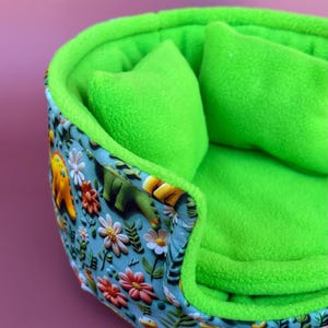 May include: A round pet bed featuring a bright green fleece interior and two matching green cushions. The exterior is covered in a light blue fabric patterned with colourful illustrations of dinosaurs, various flowers, and green foliage.