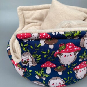 May include: A blue and white fabric hedgehog bed with a removable cushion. The bed is decorated with a pattern of hedgehogs, mushrooms, and leaves.