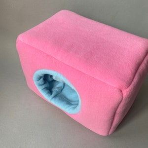 May include: A pink and blue fleece pet bed with a round entrance hole. The bed is shaped like a cube.