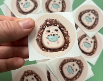 May include: A set of 10 round stickers featuring a cute cartoon hedgehog holding a cup of coffee. The hedgehog is surrounded by pink flowers and has a brown and white fur.