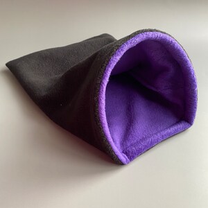 May include: A black fleece pet bed with a purple fleece lining. The bed is shaped like a tunnel and has a rounded opening.