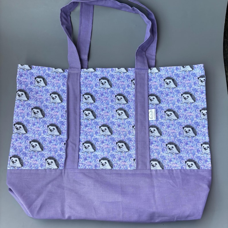 K&ouml;nnte beinhalten: Eine lilafarbene Einkaufstasche mit einem Muster aus Igeln auf helllilafarbenem Hintergrund. Die Tasche hat zwei lange lilafarbene Henkel.