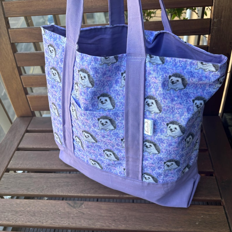 K&ouml;nnte beinhalten: Eine lilafarbene Einkaufstasche mit einem Muster aus Igeln auf helllilafarbenem Hintergrund. Die Tasche hat zwei lange Henkel und eine kleine Tasche auf der Vorderseite.