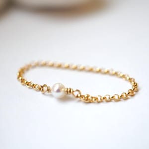 Può includere: Un delicato anello a catena dorata con una singola perla bianca e lucente al centro. Le maglie della catena dell'anello sono piccole e uniformemente distanziate, creando un aspetto raffinato ed elegante. L'anello è posto su uno sfondo bianco morbido.