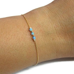 Op de afbeelding: Fijn gouden armband met drie kleine, lichtblauwe opaal kralen. De armband is dun en elegant, ontworpen om om de pols te dragen. De kralen zijn gelijkmatig verdeeld over de gouden ketting.