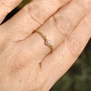 May include: A delicate gold ring with a small, round pearl set in the center. The ring band features a beaded design. The ring is displayed on a hand, with a blurred natural background.