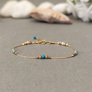 May include: A delicate gold chain bracelet with small blue and white beads. The bracelet is on a gray background.