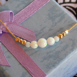 May include: A delicate gold chain necklace with five white opal beads and gold spacers. The necklace is displayed on a light blue gift box with a purple ribbon.
