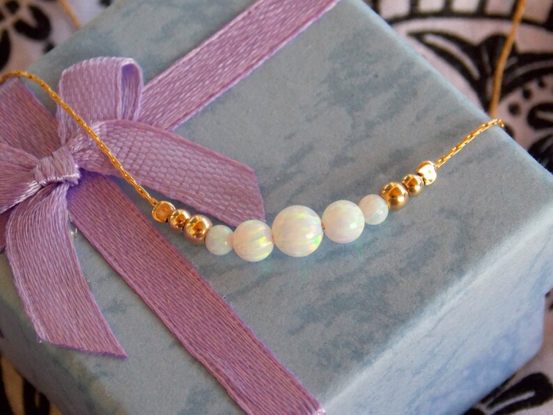 May include: A delicate gold chain necklace with five white opal beads and gold spacers. The necklace is displayed on a light blue gift box with a purple ribbon.