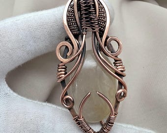 May include: A handcrafted copper wire pendant with a light-colored, oval-shaped stone at its center. The wire is intricately woven and coiled, forming a decorative frame with swirling accents. The pendant is suspended on a neutral-colored background.