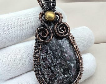 May include: A handcrafted pendant featuring a teardrop-shaped stone with a speckled pattern of black, gray, and red. The stone is intricately wrapped in copper-colored wire, with decorative swirls and a woven bail. A small gold-colored bead accents the design.
