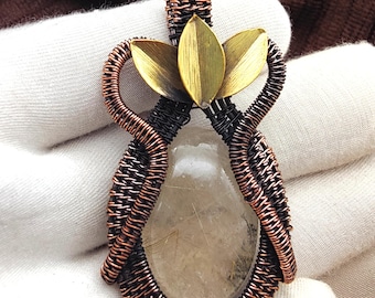 May include: A handcrafted pendant featuring a clear, oval-shaped gemstone encased in intricately woven copper and black wire. A brass lotus flower adorns the top. The pendant is held against a white fabric.