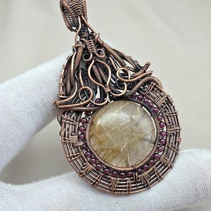May include: A handcrafted copper wire pendant with a large, round, clear quartz center. The quartz has golden inclusions and is surrounded by small, red beads. The pendant has an intricate, woven design.