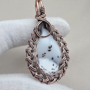 May include: A handcrafted pendant featuring a teardrop-shaped white stone with black inclusions, encased in a copper wire weave. The pendant has a copper wire bail for hanging. The overall design is intricate and artistic.