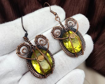 May include: A pair of handmade earrings featuring a yellow-green oval gemstone encased in a copper wire frame. The earrings have a unique design with spiral accents and a hook closure. The background is a dark brown fabric.