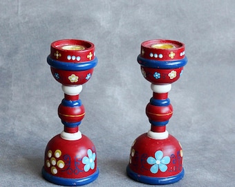 May include: Two red wooden candle holders with blue and white accents and floral designs. The candle holders have a round base, a central stem, and a top cup to hold a candle. The floral designs include blue and yellow flowers.