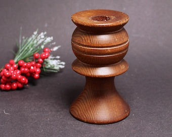 May include: A wooden candle holder with a dark brown finish. The candle holder has a round base, a cylindrical middle section with horizontal grooves, and a top with a central hole. A sprig of red berries and greenery is in the background.