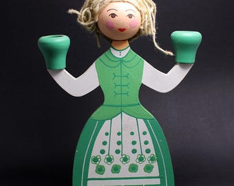 Puede incluir: Figurita de madera de una mujer con un vestido verde con detalles blancos, sosteniendo dos candelabros verdes. La figurita tiene pelo rubio de hilo, una cara pintada y una base verde. Un artículo decorativo.