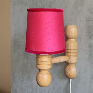 Può includere: Una lampada da parete con un paralume fucsia vibrante e una struttura in legno. Il paralume ha un bordo in velluto. La lampada è fissata a un muro grigio con un cavo di alimentazione bianco.