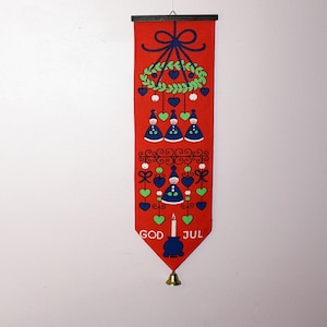 Puede incluir: Una pancarta de tela roja con un tema navideño. Presenta una corona verde con un lazo azul, tres figuras azules, una vela y el texto "God Jul".