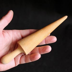 May include: A light-colored wooden rolling pin with a conical shape, held in a hand against a black background. The rolling pin has a handle and a tapered body, ideal for shaping dough or clay.