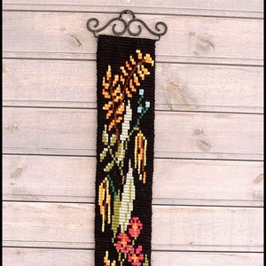 Könnte beinhalten: Eine schwarze Wandteppich mit einem floralen Design mit gelben, orangefarbenen, grünen, rosa und blauen Blumen. Der Wandteppich hängt an einer Holzwand mit einem schwarzen Metallhaken.