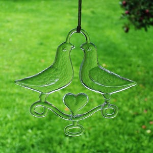 Puede incluir: Un adorno de vidrio con dos palomas con un coraz&oacute;n en el centro, colgado de una cuerda marr&oacute;n.
