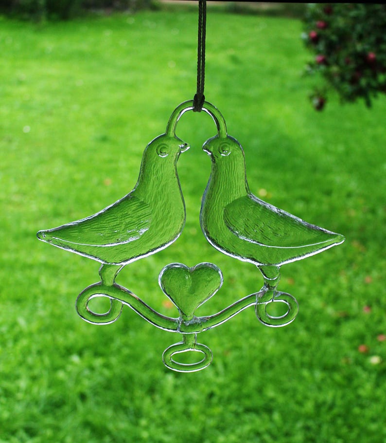 Puede incluir: Un adorno de vidrio con dos palomas con un coraz&oacute;n en el centro, colgado de una cuerda marr&oacute;n.
