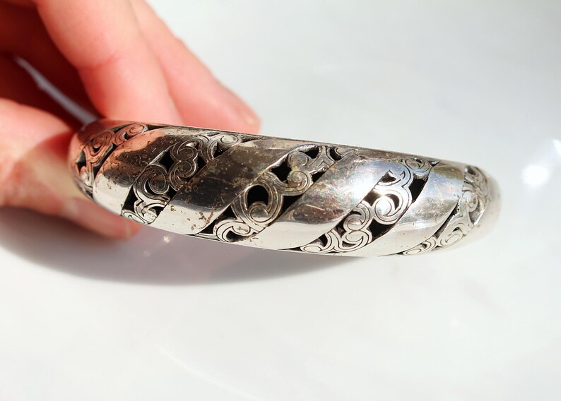 Puede incluir: Un brazalete plateado con un dise&ntilde;o calado. El brazalete presenta bandas entrelazadas con patrones arremolinados. El metal tiene un aspecto ligeramente envejecido. El dise&ntilde;o del brazalete recuerda a los nudos celtas.