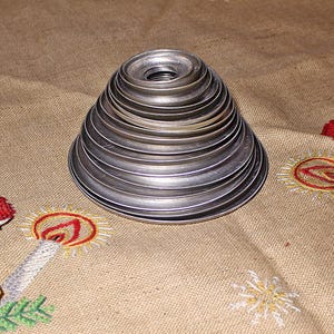 Puede incluir: Una pila de anillos de metal plateado, de tama&ntilde;o decreciente, que forman una forma c&oacute;nica. El objeto est&aacute; sobre una tela beige con dise&ntilde;os bordados de velas navide&ntilde;as y Pap&aacute; Noel.
