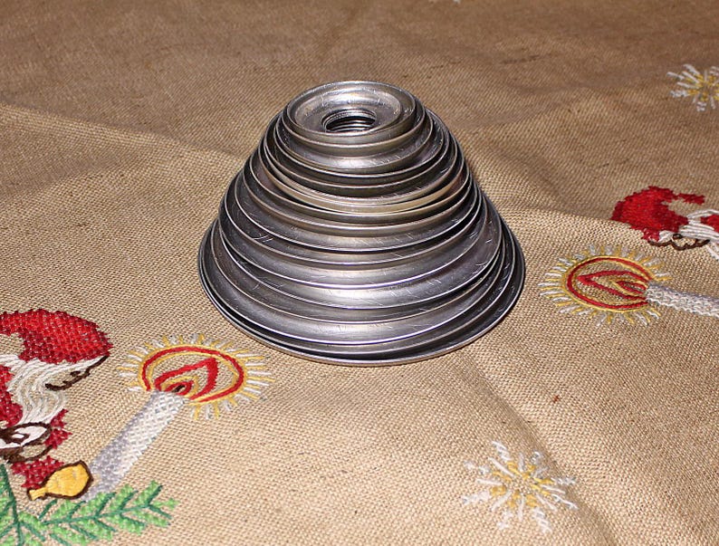 Puede incluir: Una pila de anillos de metal plateado, de tama&ntilde;o decreciente, que forman una forma c&oacute;nica. El objeto est&aacute; sobre una tela beige con dise&ntilde;os bordados de velas navide&ntilde;as y Pap&aacute; Noel.